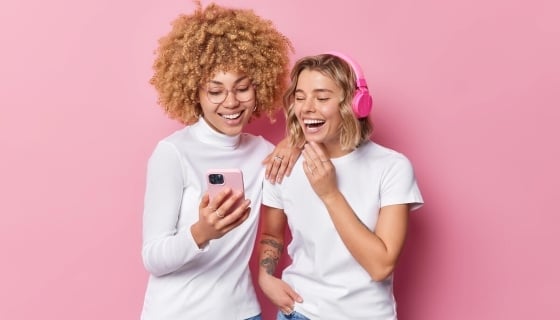 joyeuses-jeunes-femmes-joyeuses-regardent-video-amusante-smartphone-moderne-s-amusent-ecouter-musique-via-ecouteurs-vetus-vetements-decontractes-posent-fond-rose-concept-personnes-technologies-m